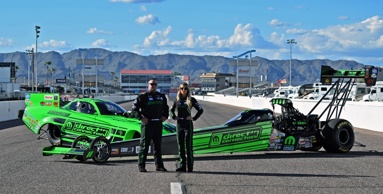 An image of a man and a woman in front of m direct connection wrapped race cars on a race track