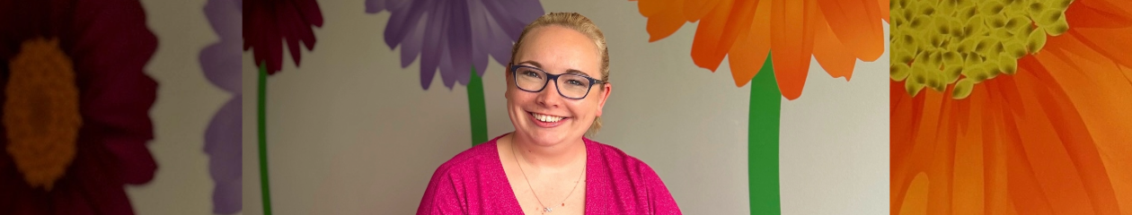 A woman with blonde hair and glasses sits casually on an orange couch, smiling at the camera