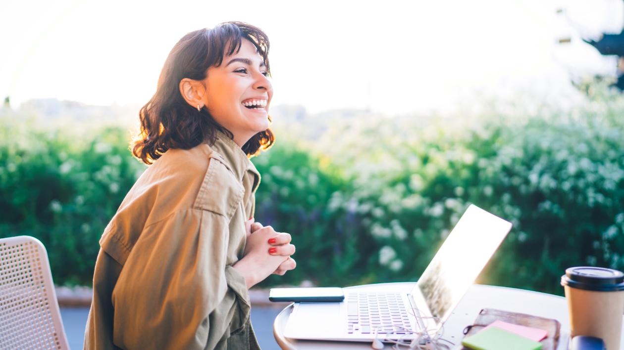 A woman sits and smiles at an outdoor patio, surrounded by her laptop, other work materials and a cardboard coffee cup